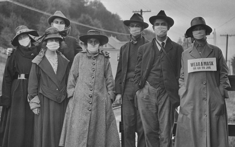 Black and white photograph of a group of people standing outside in coats and hats wearing masks. One holds a sign that says 