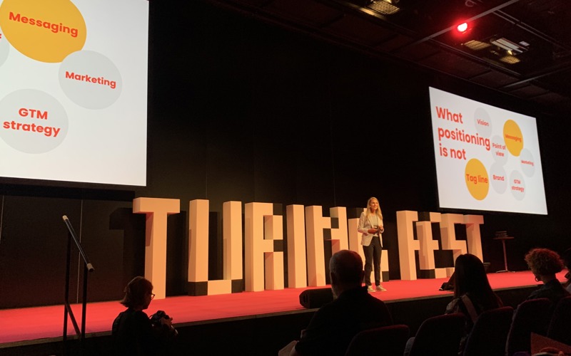 A person stands on a stage, speaking to a crowd. On the stage are a large sculptured letters reading 