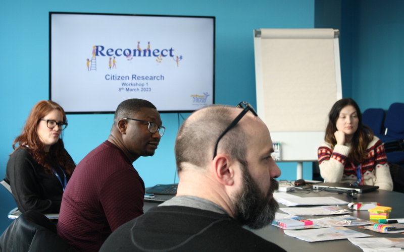 A group of people sit around a table looking at someone off-camera. A screen in the background displays the words 