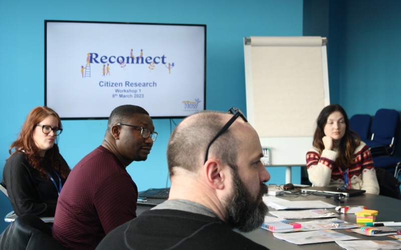 A group of people sit around a table looking at someone off-camera. A screen in the background displays the words 