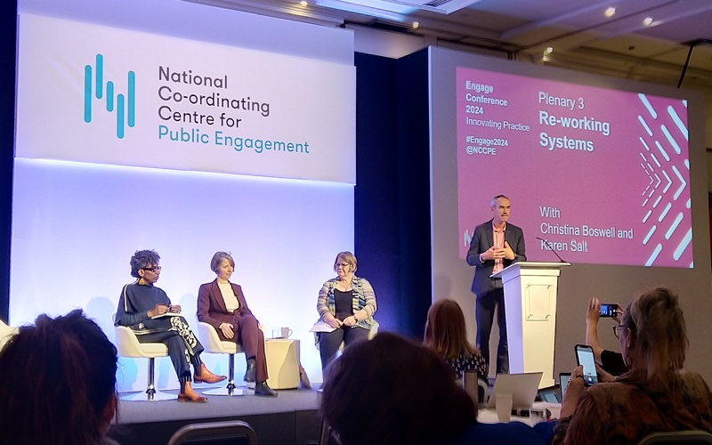 Four people on a stage. One of them is presenting to a room of people.