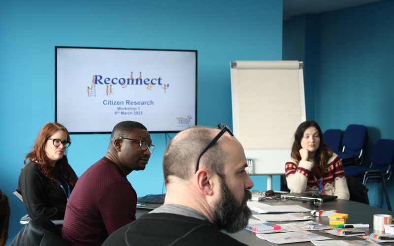 A group of people sit around a table looking at someone off-camera. A screen in the background displays the words 