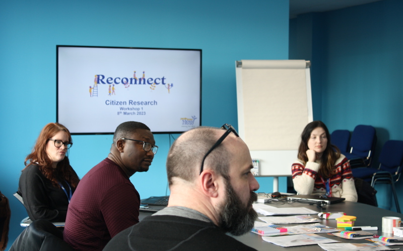 A group of people sit around a table looking at someone off-camera. A screen in the background displays the words 