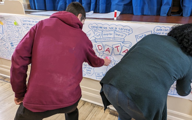 Two people facing away from the camera are sticking post-it notes onto a long sheet of paper. The paper has notes from a discussion about data written and illustrated on it