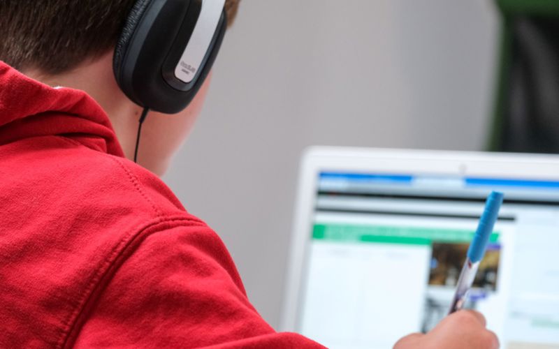 A young person in a red hoodie with headphones on faces away from the camera, looking at a laptop screen.