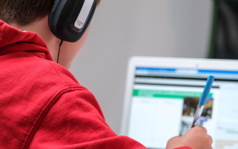 A young person in a red hoodie with headphones on faces away from the camera, looking at a laptop screen.